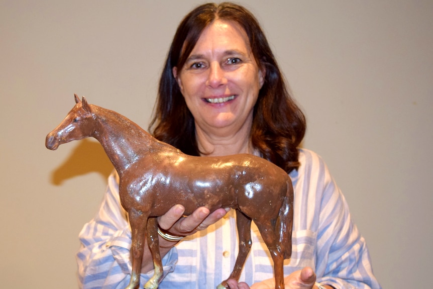 A dark-haired smiling woman holds a ceramic brown horse