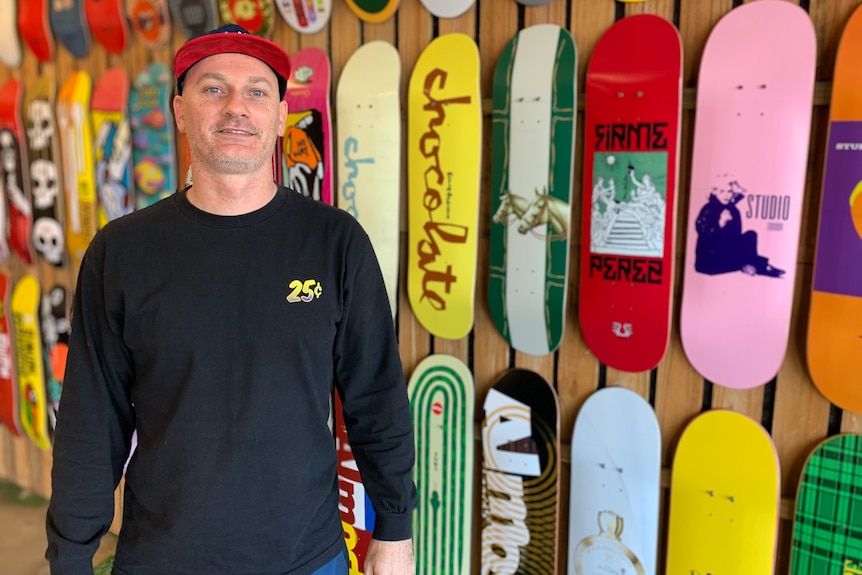 Man standing in front of wall filled with skateboards. He is wearing a black shirt and red cap