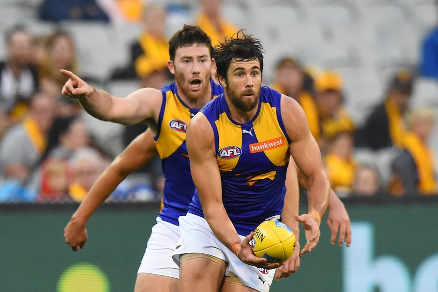 West Coast's Josh Kennedy carries a yellow football as teammate Jeremy McGovern points the way behind him.