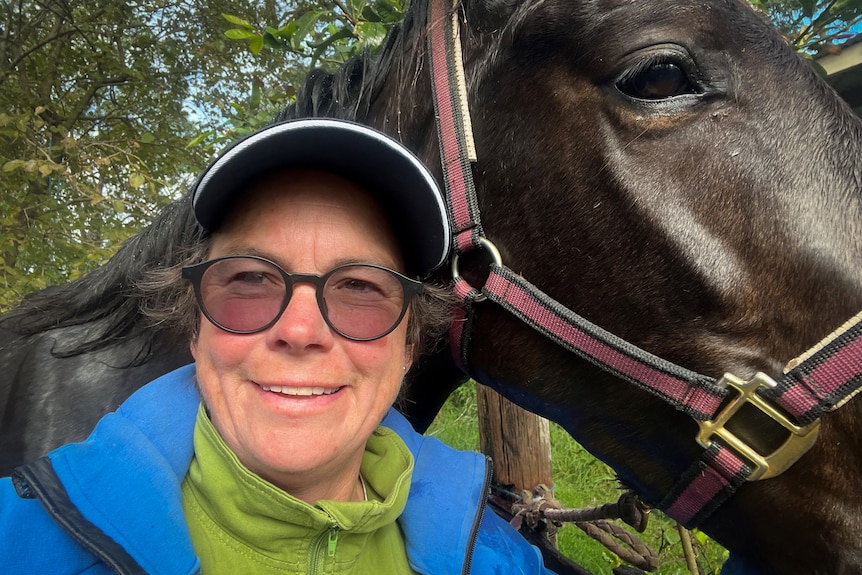 Close up shot of a woman in a cap and tinted glasses standing close to a horse, smiling