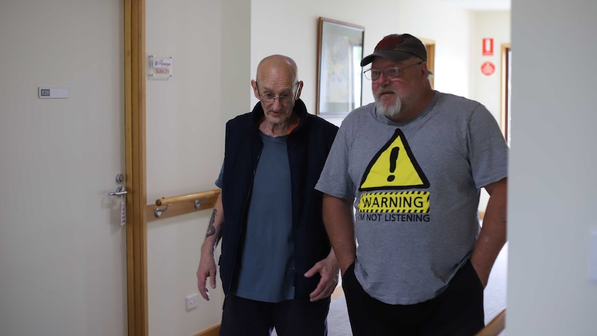 Two older men standing next to each other, walking down a nursing home corridor.