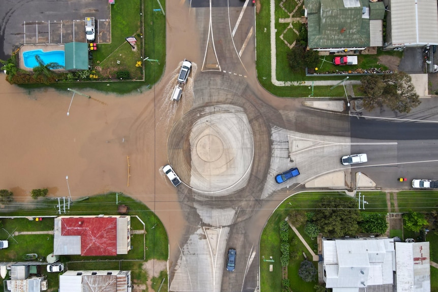 Cars driving around a roundabout with brown floodwater lapping at the edges.