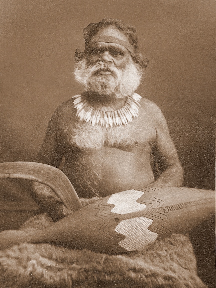 An indigenous man sits with traditional weapons and necklace.