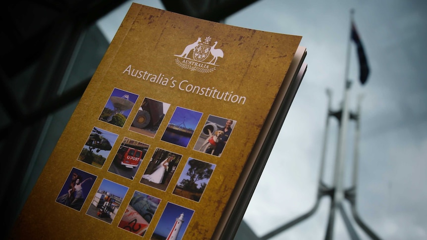 A book titled Australia's Constitution held in front of a window. Outside, the Parliament House flag flies against a stormy sky.