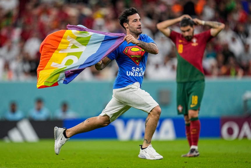 A man running across a field flying a rainbow flag with a soccer player in the background
