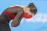 A figure skater wearing black and red puts her head in her hands after falling during a routine
