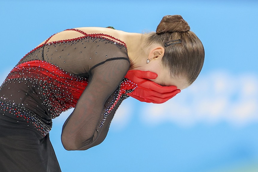 A figure skater wearing black and red puts her head in her hands after falling during a routine