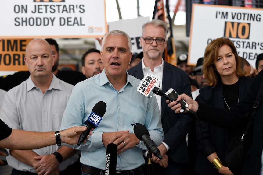 A middle-aged man talks to microphones with people behind him listening and holding placards.