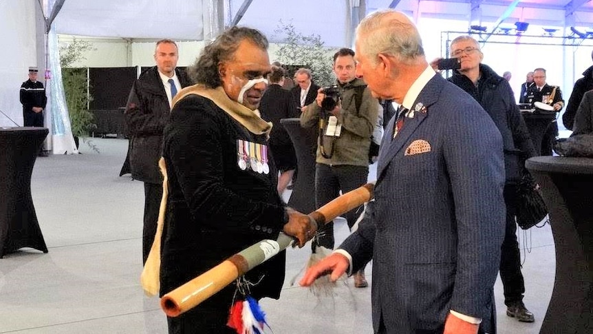 An Indigenous man holding a didgeridoo and wearing war medals speaks with Prince Charles, in a dark blue suit.