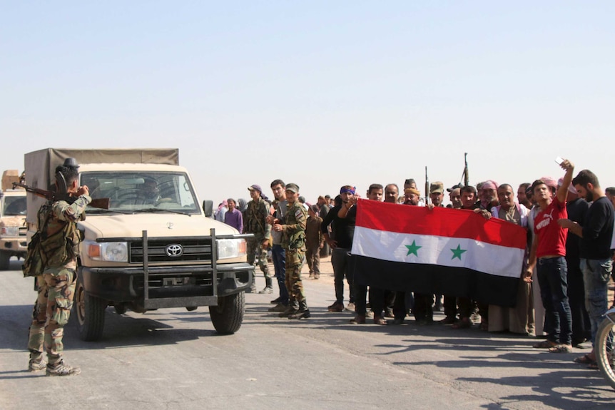A group of men hold up a flag while standing next to an army vehicle and soldiers