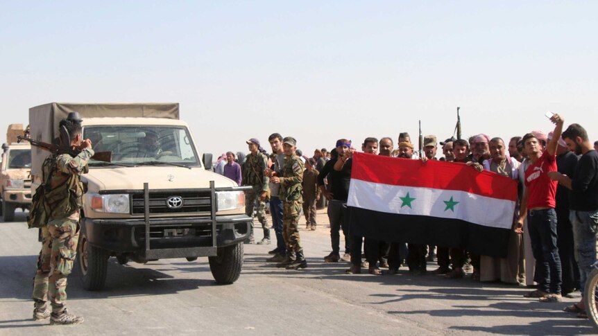 A group of men hold up a flag while standing next to an army vehicle and soldiers