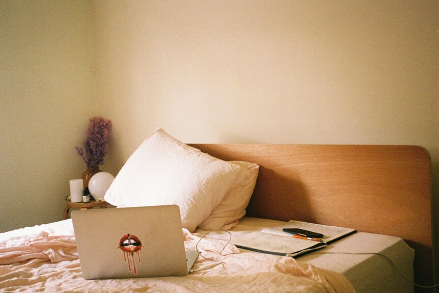 An unmade double bed with note pads, an open laptop and propped-up pillows