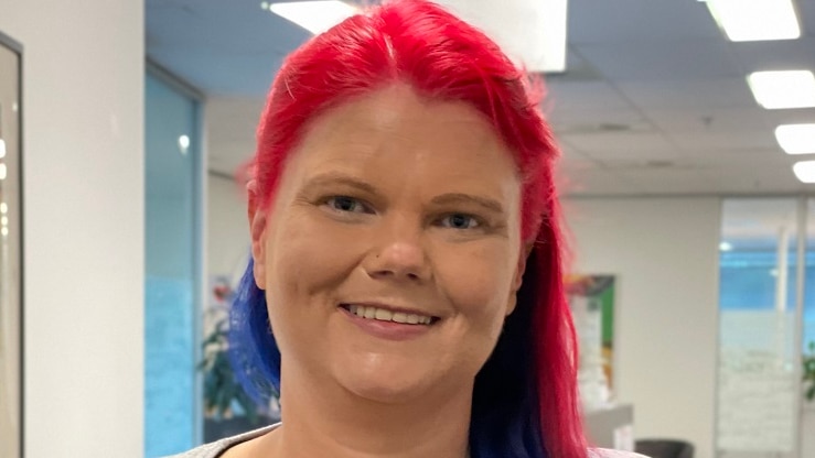 A woman with pink and blue hair smiles gently, standing in an office corridor.