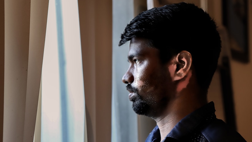 Man with skin visibly affected by burns on cheek and ear stares out of window, with light coming through blinds.