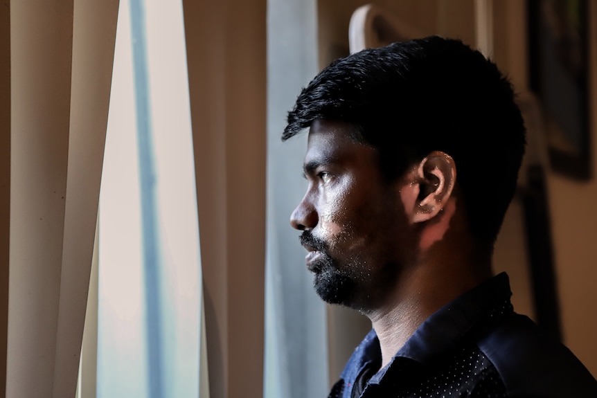 Man with skin visibly affected by burns on cheek and ear stares out of window, with light coming through blinds.