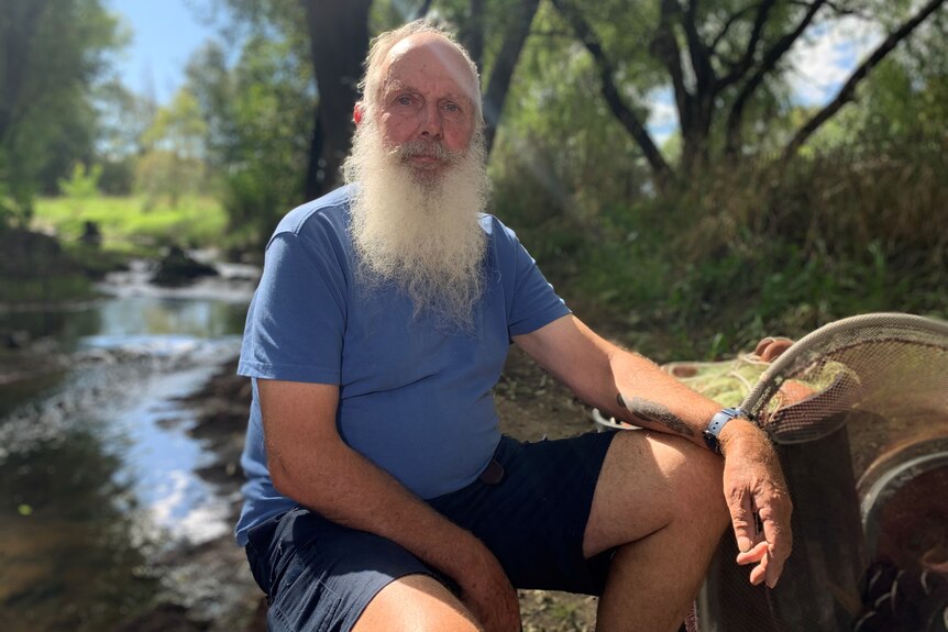 A man with a grey beard sits next to a small stream
