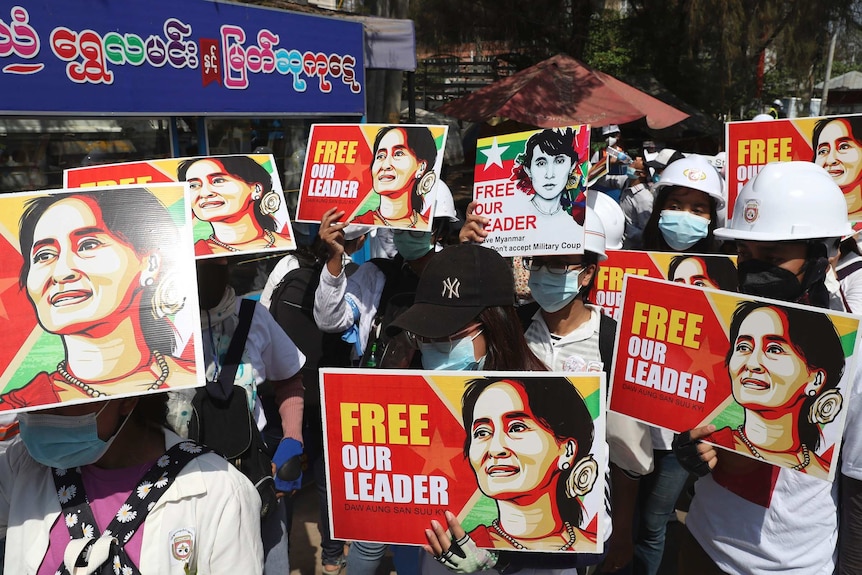 Protesters hold images of deposed Myanmar leader Aung San Suu Kyi as they march along a street in the sunlight.