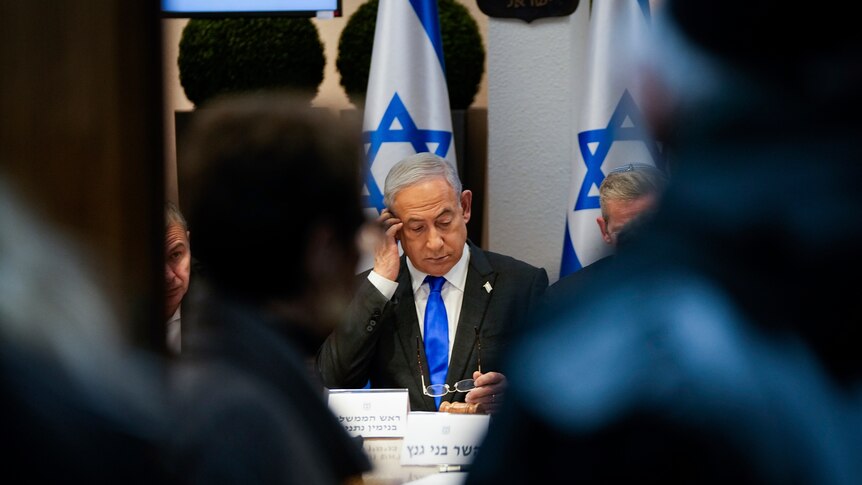 Benjamin Netanyahu sits at table with people either side. He is looking down. Behind him are Israeli flags.