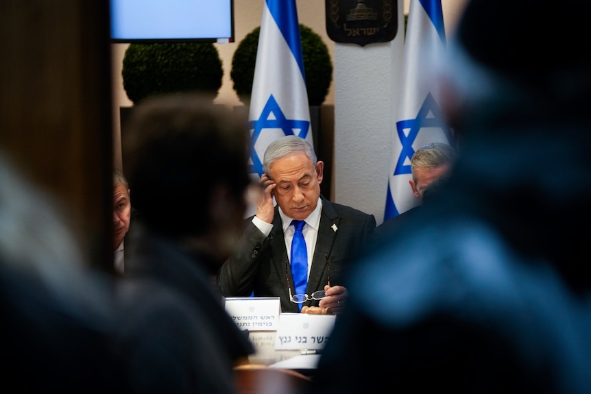 Benjamin Netanyahu sits at table with people either side. He is looking down. Behind him are Israeli flags.
