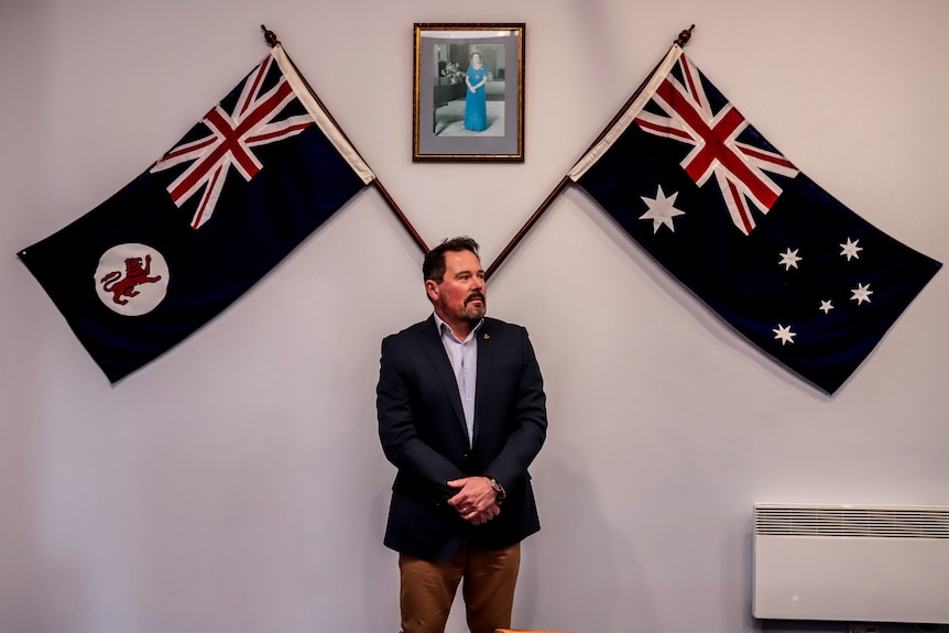 Man with brown hair standing wearing suit beneath two large Australian flags and a picture of the Queen on a wall