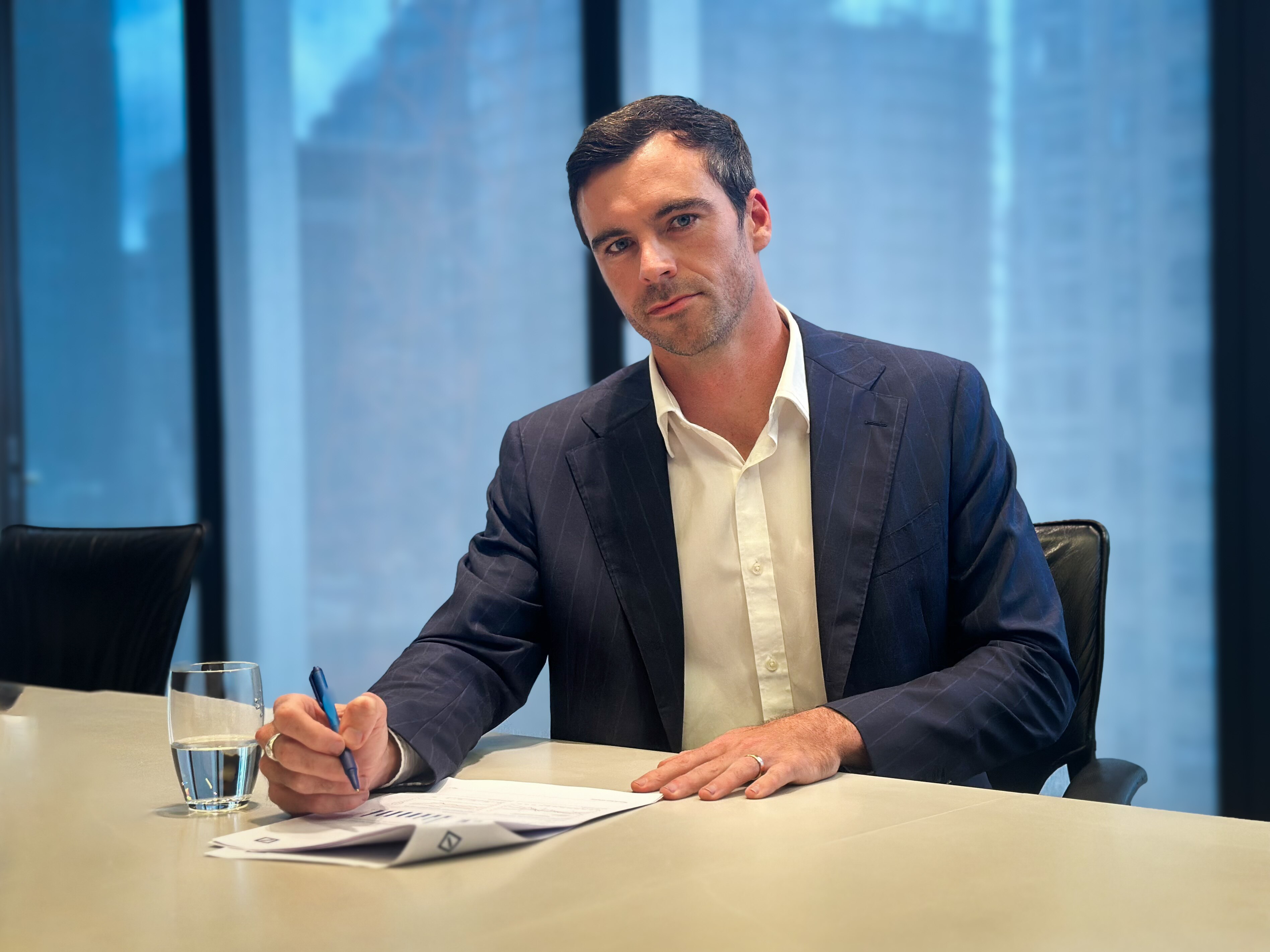 Lachlan, looking serious, sits at his desk with a pen and document.