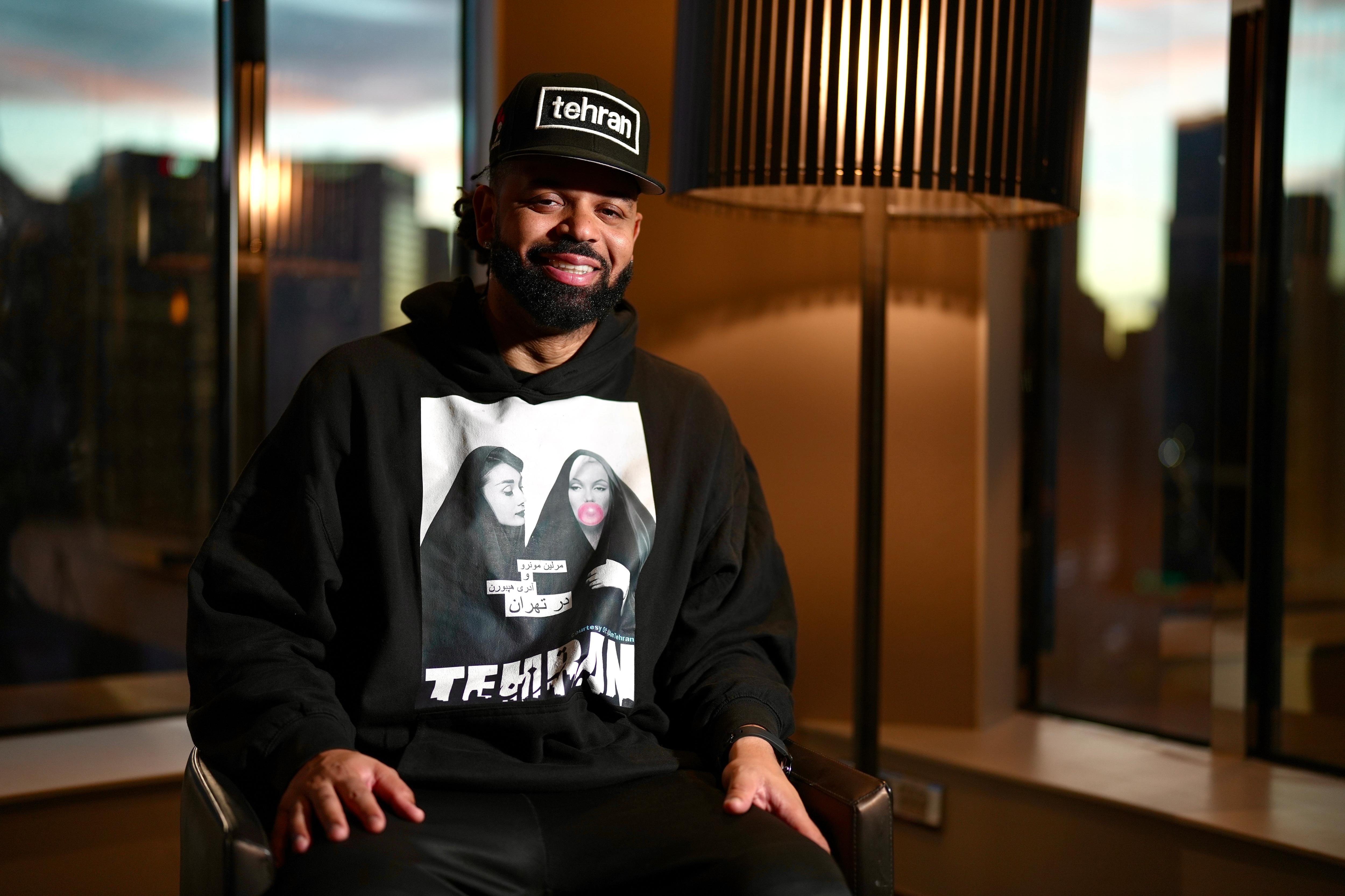 A man in a black sweater and baseball cap siting in a  hotel smiling 