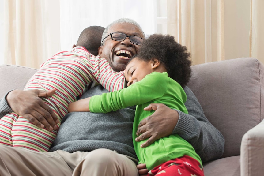 Older man smiles on couch hugged by two kids