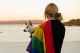 A person holding a dog on a beach, a rainbow flag draped over one shoulder.