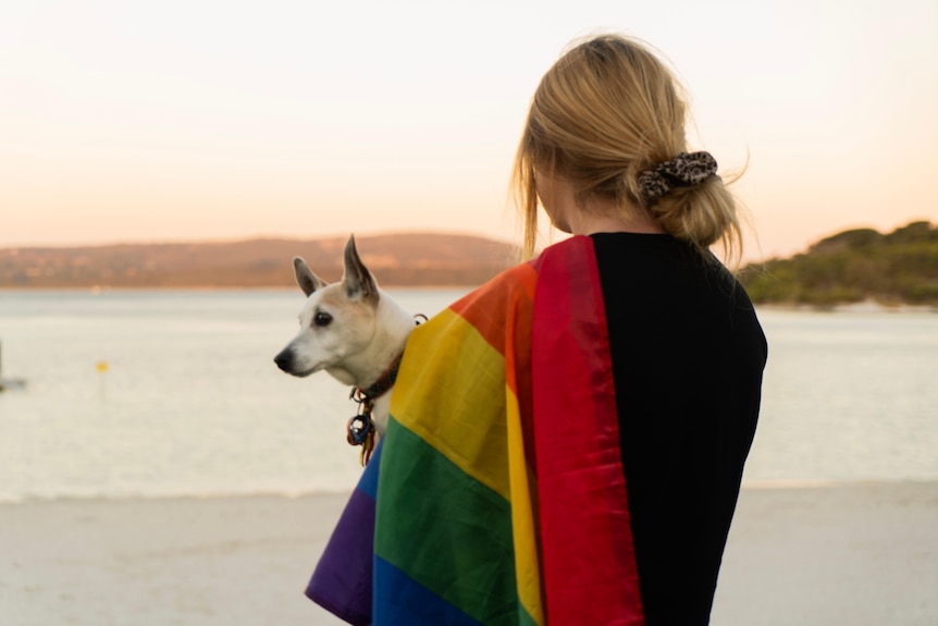 A person holding a dog on a beach, a rainbow flag draped over one shoulder.