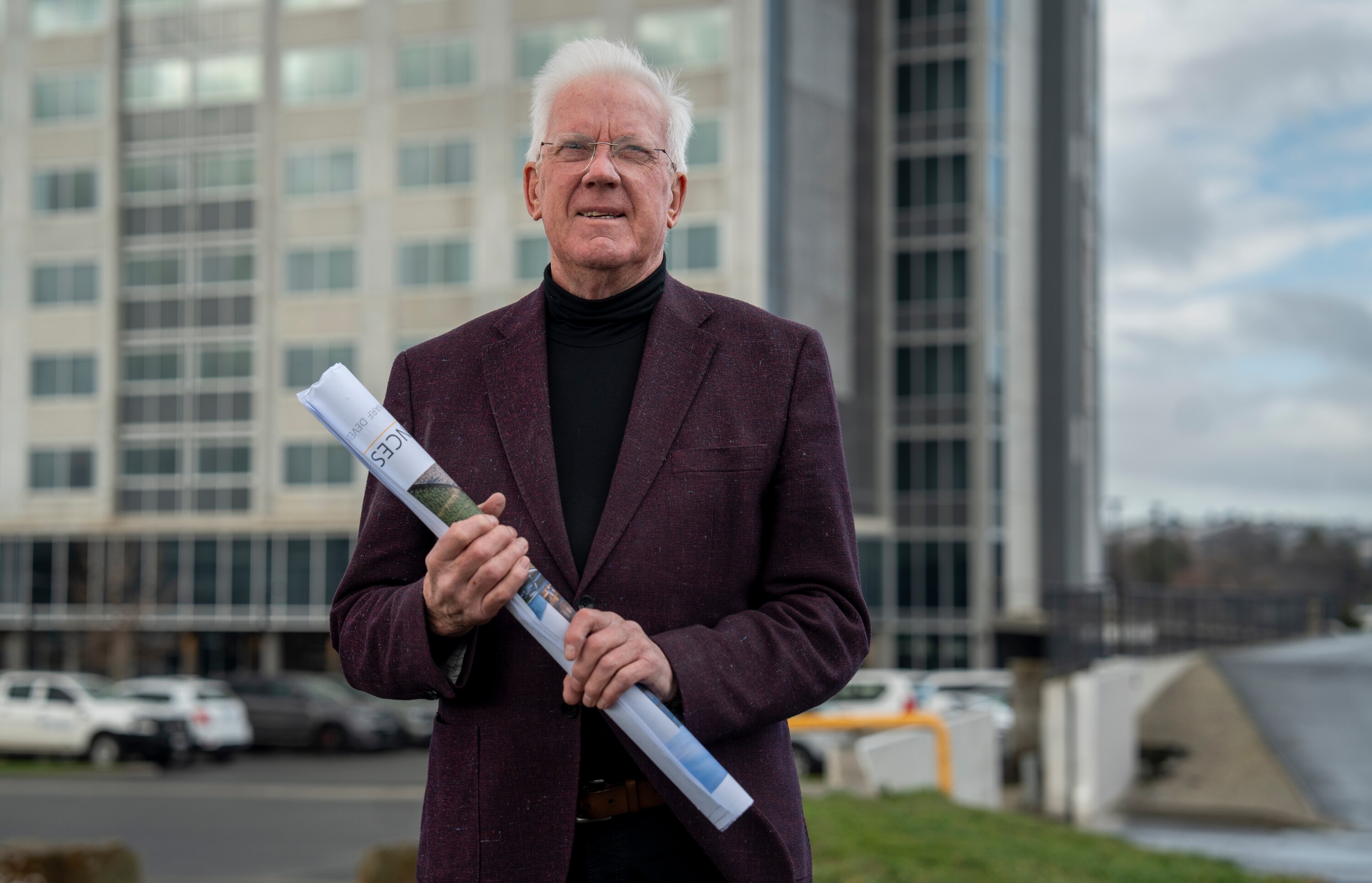 A gentleman in a maroon blazer and glasses holds a rolled up building plan outside of a rectangular grey building.