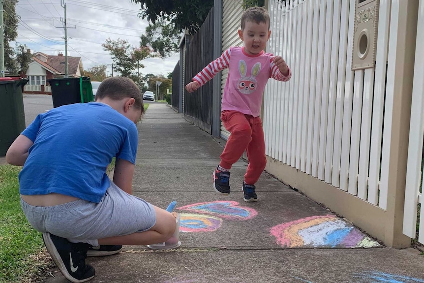 Two boys on the footpath, one drawing with chalk, the other skipping by.