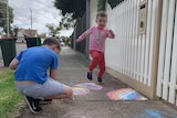Two boys on the footpath, one drawing with chalk, the other skipping by.