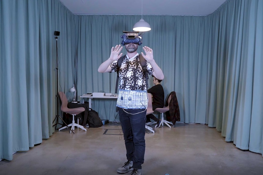 A man with a VR headset and haptic belt on in a room with curtains in the artwork Breakout My Pelvic Sorcery