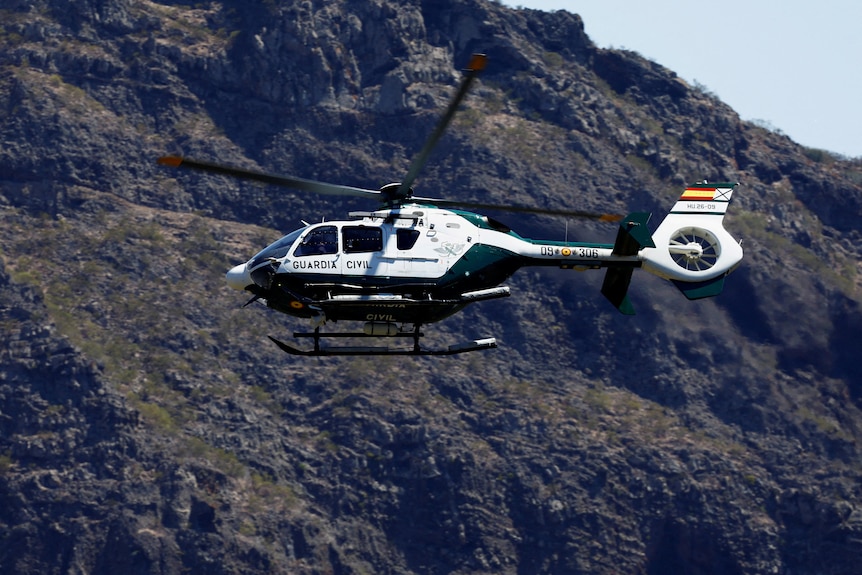A white and green helicopter flies through the sky with a rocky outcrop behind it