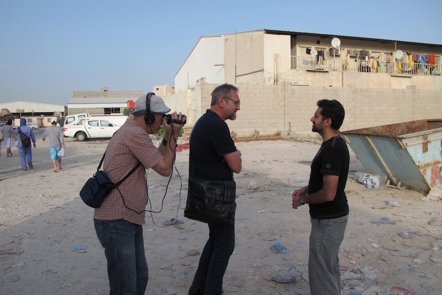 Two men speak to each other while a cameraman films them. A cement house behind, rubbish on dirt ground. 