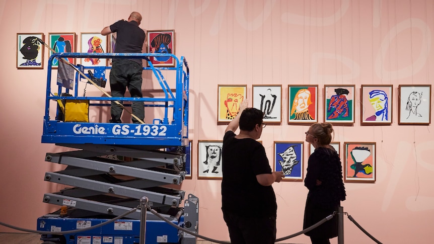 The artist points to his paintings as a man in an elevated machine hangs them on the wall.