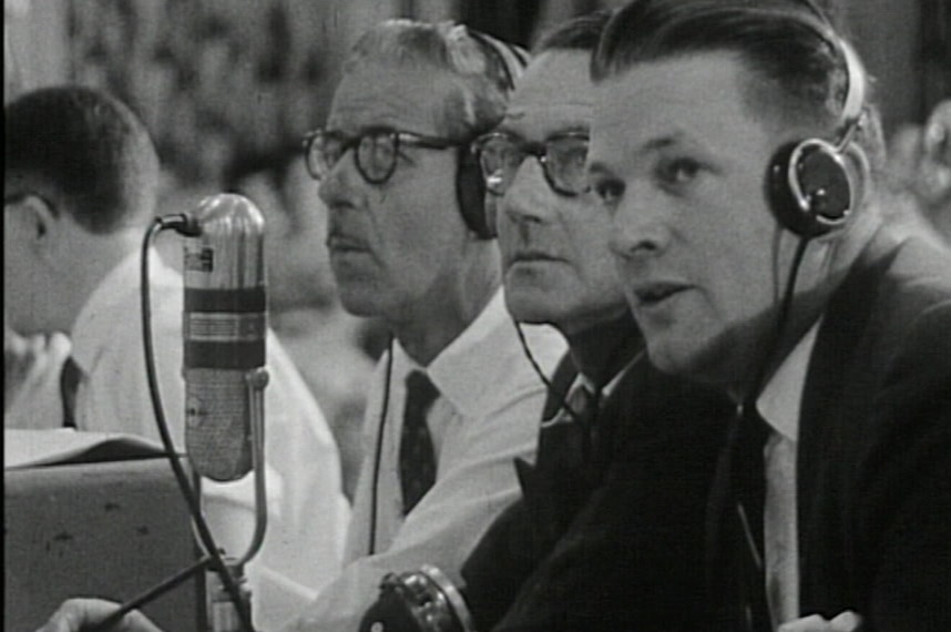Black and white still of announcers wearing headphones talking into old fashioned microphone in tally room.