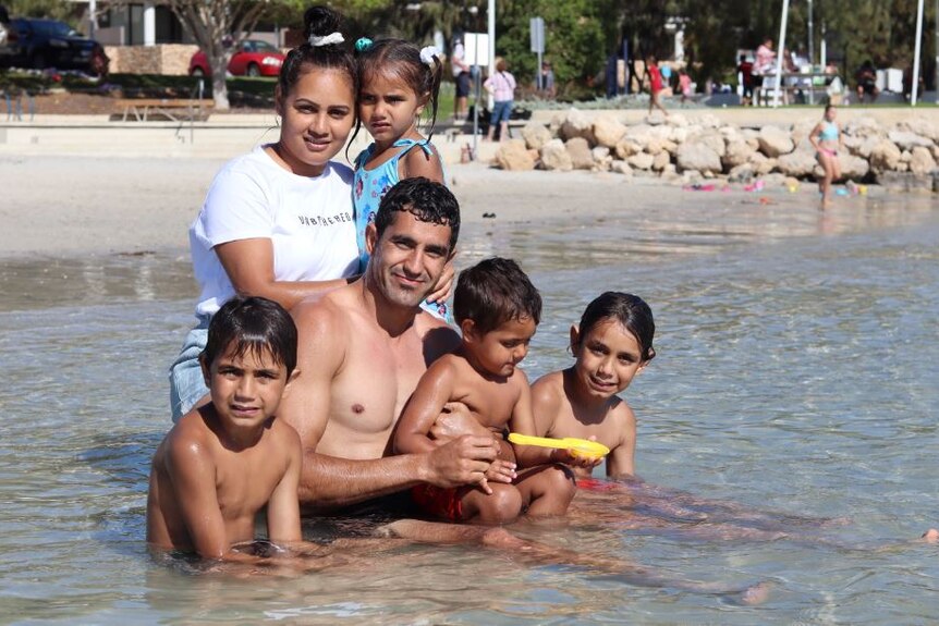 A man and a woman and four children sit in shallow water at the beach