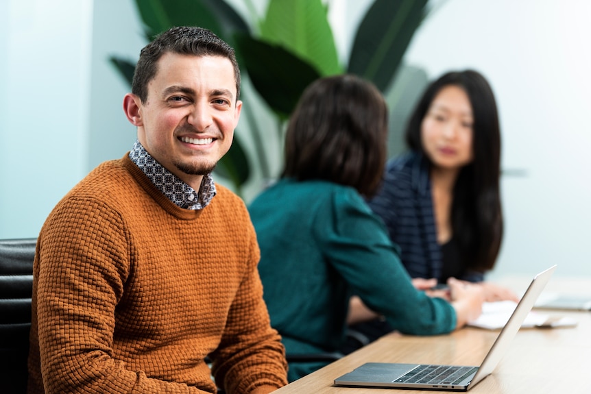 A man in a brown jumper sitting next to a laptop and in front of two women talking. 