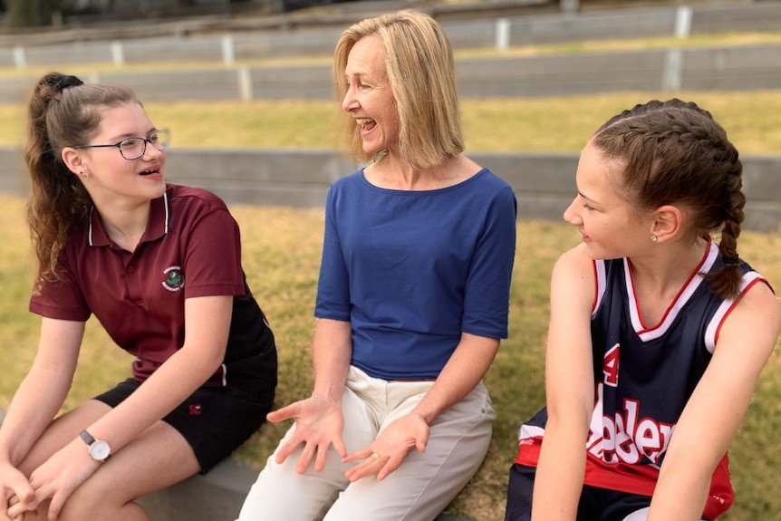 Two school girls wearing sports uniforms listen to a woman speaking.