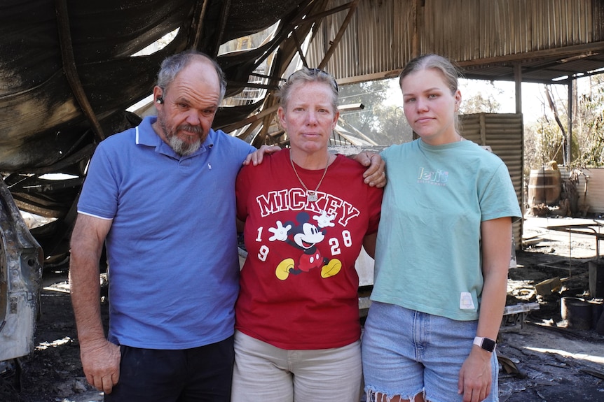 A family of three stand with their arms around each other's shoulders amid a charred shed