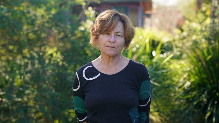 A woman stands and looks at the camera in front of a bush