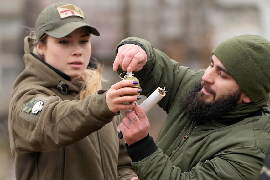 A woman holds a hand grenade at arm's length as a man shows her how to pull the pin