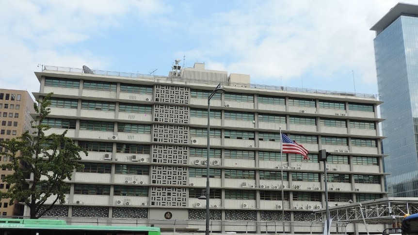Shot of the US Embassy in Seoul, a broad beige building, with the American flag flying, and traffic in front