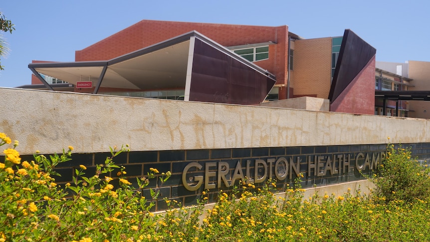 Flowers in the foreground of a sign reading Geraldton Health Campus, with the backdrop of an angular two storey hospital. 