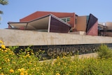 Flowers in the foreground of a sign reading Geraldton Health Campus, with the backdrop of an angular two storey hospital. 