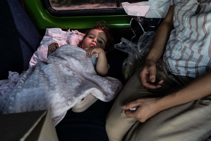 A baby with a blanket draped over it sits on the backseat of a car.