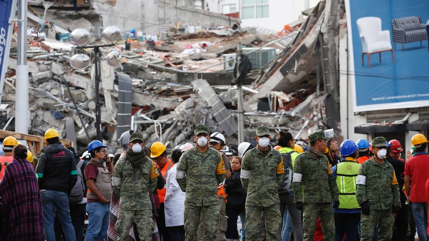 Dozens of soldiers wearing hard hats and surgical masks stand next to a collapsed building.