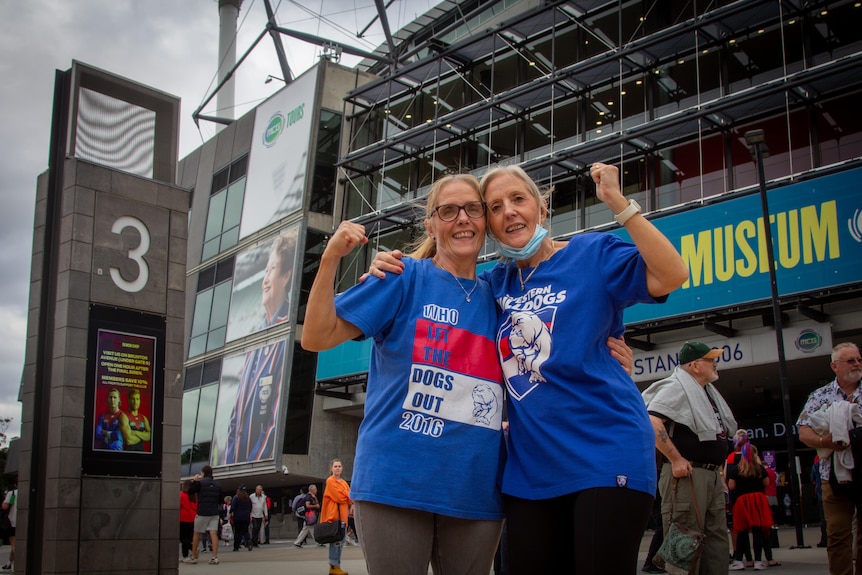 Two middle age women stand in front of the MCG cheering wearing Western Bulldogs colours