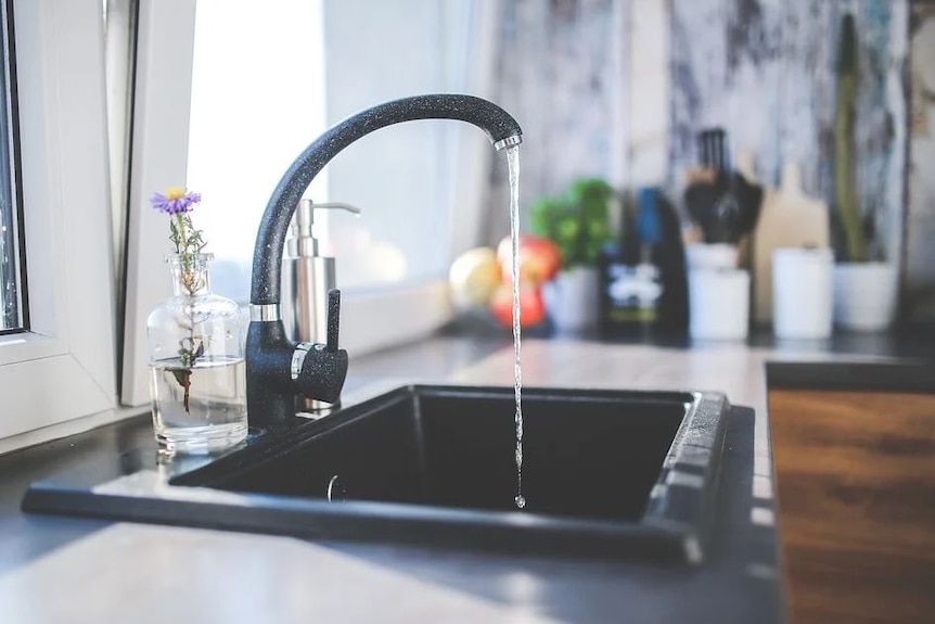 Gray kitchen sink and faucet with running water, glass vase with flower sits on the bench